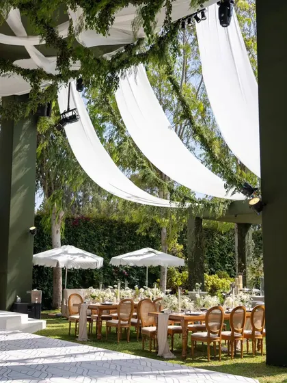 A view from under the custom-built green pergola, showing how the draped white fabric creates a soft, shaded area for guest seating. This structure adds architectural interest to the garden setting.