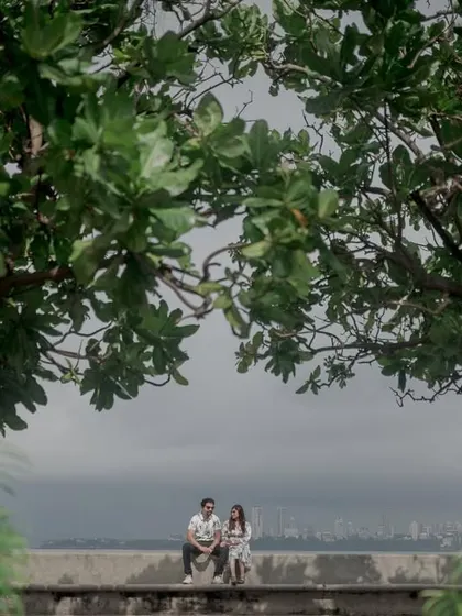 A serene, wide shot of a couple sitting at Marine Drive, framed by lush green trees. It captures a peaceful moment amidst the bustling city.