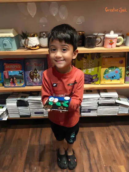 A smiling young boy holds his finished clay project, a colorful scene with birds on a branch, created during a kids' workshop.