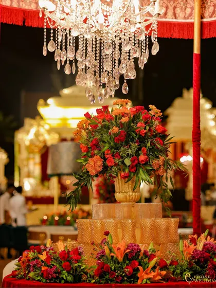 A beautiful centerpiece featuring a crystal chandelier suspended over a lush floral arrangement. This detail shot highlights the layers of opulence, from the sparkling crystals to the rich red and orange blooms.