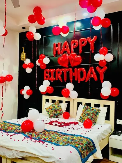 A classic romantic birthday bedroom setup. The decor features red and white balloons, "Happy Birthday" foil letters, and rose petals on the bed, perfect for surprising your partner.