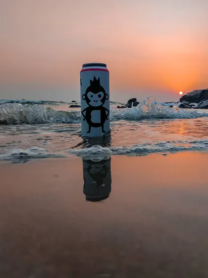 A beverage can with a monkey logo sits on a beach at sunset, its reflection captured in the wet sand. This is a great example of lifestyle product photography, placing the product in an appealing context.