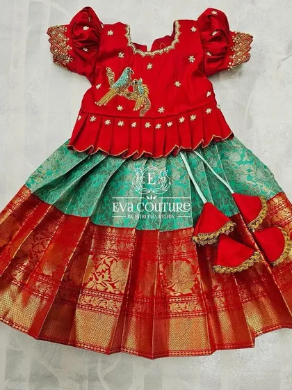 A beautiful red and green pattu pavadai for a baby. The red blouse has a small embroidered bird motif, and the skirt has a traditional red and gold border.