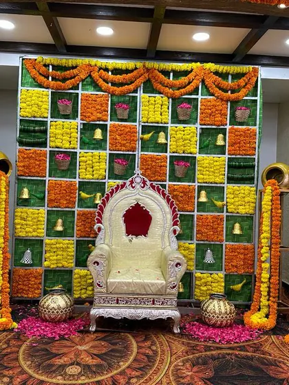 A traditional ceremony stage with a checkered backdrop of marigolds and green panels, flanked by decorative pots.