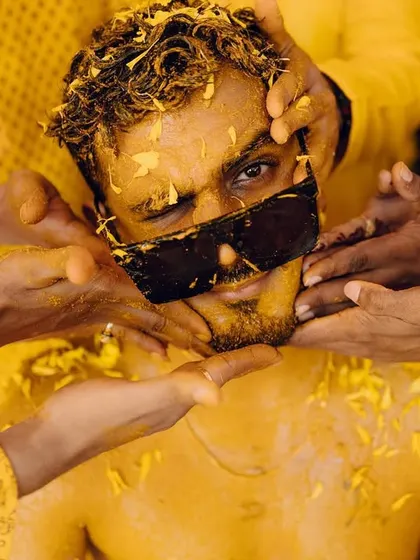 A fun, close-up shot of the groom being playfully covered in Haldi by his friends. His expression, seen through his sunglasses, is one of pure amusement.