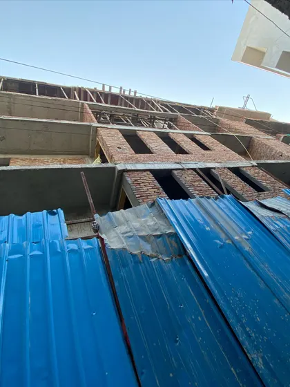 A low-angle view of a modern house under construction in New Delhi. The image shows the raw brick and concrete structure rising behind the site barrier.