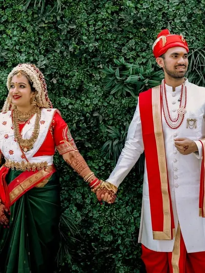 A full-length portrait of a traditional Marathi couple against a green wall. Their coordinated red, white, and green outfits create a visually stunning and culturally rich image.