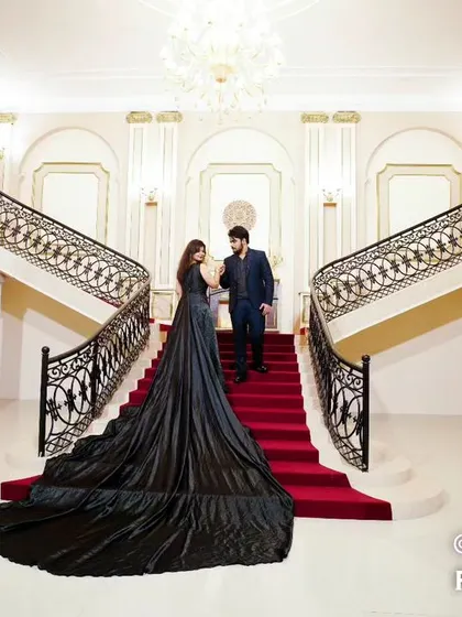 A wide shot of a couple on a grand staircase with a red carpet, her black trail gown spread out, creating a dramatic and cinematic entrance scene.