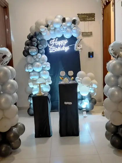 A sleek and modern setup for a 4th birthday using a black, silver, and white color scheme. The decor includes a balloon arch, matching balloon pillars with heart-shaped foil balloons, and a bright neon sign.