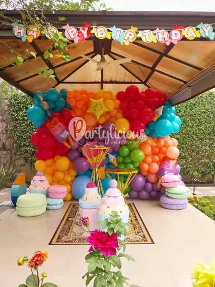 Welcome to Candyland. This incredibly colorful setup features a rainbow balloon wall, giant macaron props, and cupcake stands, creating a sugar-filled wonderland.
