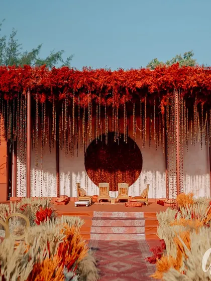 The wedding ceremony stage, set up for the pheras. The design is grand yet intimate, with a canopy of red florals and hanging elements over a beautifully detailed seating area.