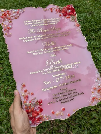 This geode-shaped resin wedding invitation in pink is adorned with natural dried flowers and rose quartz crystals, which represent love and bonding. The details are printed in golden metal stickers for a truly royal feel.