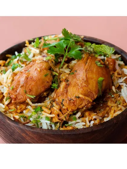A close-up shot of chicken biryani in a rustic wooden bowl against a dusty rose background. The tight crop highlights the texture of the rice, chicken, and fresh herbs.