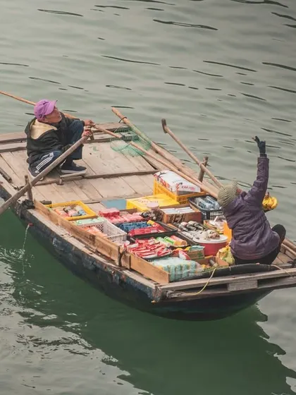 Two vendors in a small boat in Halong Bay, with one waving up towards the camera on a cruise ship.