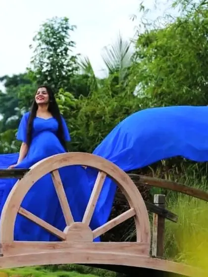 A maternity client poses on a small wooden bridge, with the trail of her blue flying dress flowing behind her.