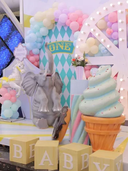 A close-up of the pastel carnival stage, showing the giant ice cream prop, a playful elephant, and "BABY" blocks. The soft colors and whimsical elements are perfect for a first birthday.