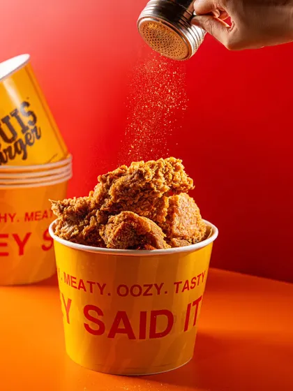 A bucket of fried chicken from Louis Burger with seasoning being sprinkled on top. This action shot adds a dynamic and appetizing element to the product photo.