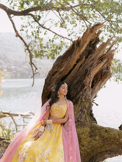 The bride in her bright yellow lehenga finds a moment of peace, posing against a dramatic, ancient tree by the lakeside.