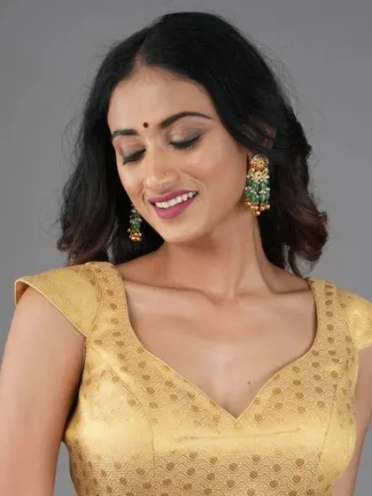 A model wears a gold brocade blouse with cap sleeves, accessorized with large green and gold statement earrings.