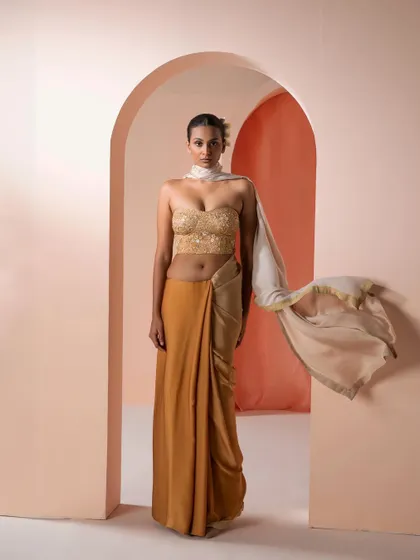 A full-length shot of a model in a golden draped gown with a corset top. The flowing fabric and architectural background create a sense of modern artistry.
