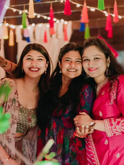 Friends and sisters celebrating together. The colorful tassel decor and warm lighting in the background create a festive and joyful atmosphere for this mehendi night.