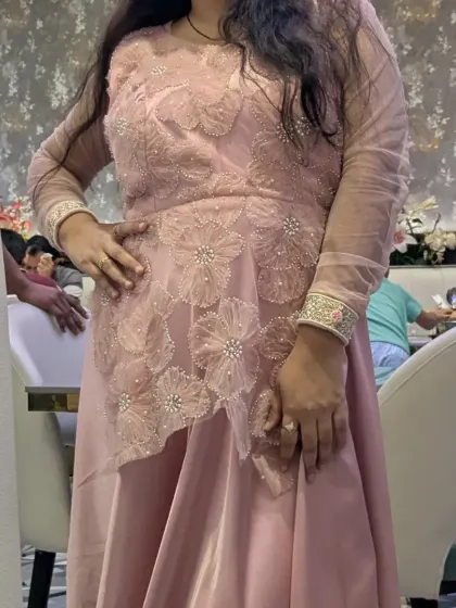 This decent pastel pink party wear gown features a unique asymmetrical floral overlay. It's a comfortable and stylish option available for rent.