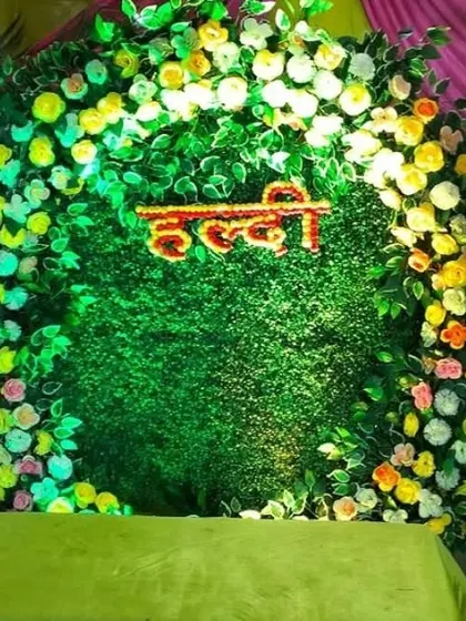 A lush green Haldi backdrop made of an artificial grass wall, framed by a circle of colorful flowers. The 'Haldi' sign is made from red flowers for a striking contrast.