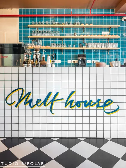 The service counter at Melthouse cafe features the brand's logo in a flowing script on a clean, white-tiled facade. The classic black-and-white checkered floor and turquoise back-tiling create a retro, welcoming feel.
