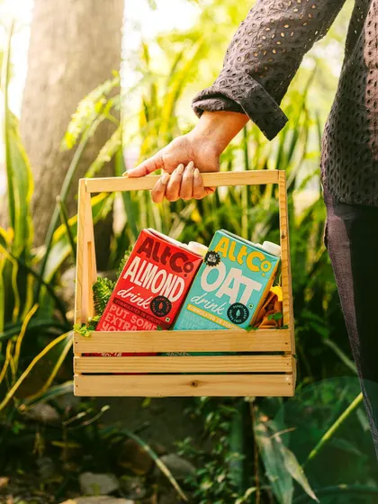 A lifestyle shot of a person carrying a basket of Alt Co.'s almond and oat milks through a sunlit garden. This image evokes a fresh, farmers-market feel, associating the brand with natural goodness.