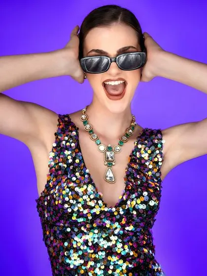 Fun, energetic, and full of life. The combination of a sequined dress, sunglasses, and a joyful expression against a solid purple background creates a dynamic and playful party look.