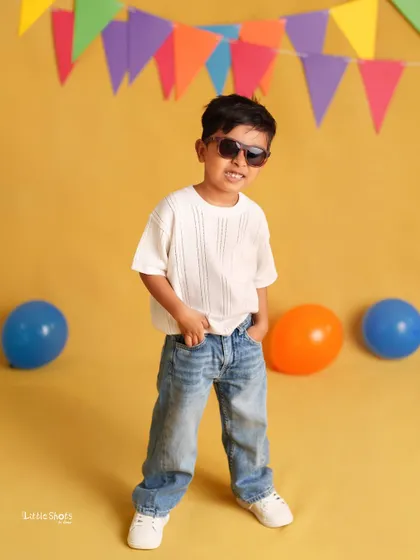 This cool little guy is rocking his sunglasses and casual jeans for his birthday shoot. The colorful party flags and bright yellow background add to the fun vibe.