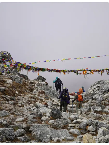 Trekkers walking along a rocky path lined with prayer flags, with misty clouds obscuring the valley below. The weather in the Himalayas is unpredictable, adding to the adventure.