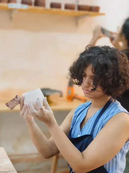 A student inspects her white stoneware mug, carefully checking the form and handle attachment. We teach the structural principles needed to create durable and functional handmade pottery.