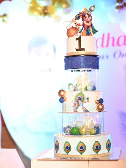 Our grand Krishna-themed cake for a first birthday. This multi-tier design features clear separators filled with lights and balls, peacock feather details, and a beautiful Krishna topper.