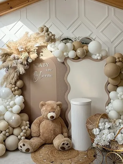 A "Baby in Bloom" themed baby shower backdrop. This setup features a large teddy bear, a beautiful balloon arch in neutral tones, pampas grass, and a vintage-style pram filled with white flowers for a boho-chic look.