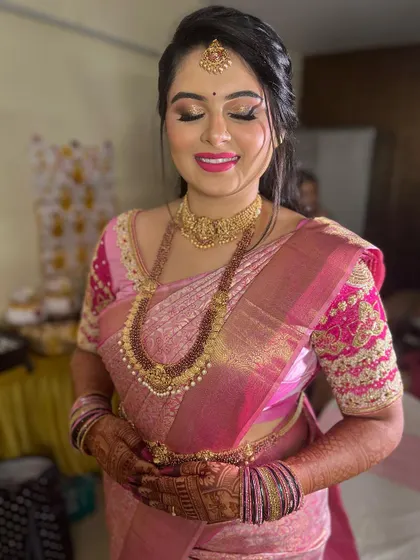 This bride is glowing with happiness. Her makeup features a flawless base, a subtle shimmer on the eyes, and a bright pink lip that perfectly matches her beautiful pink silk saree and temple jewellery.