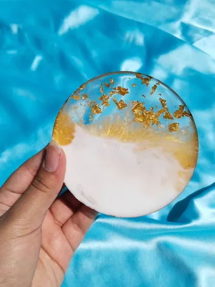 A round resin coaster featuring a beautiful ocean-inspired design. The piece blends white and translucent blue resin, with gold flakes scattered along the edge for a touch of luxury.