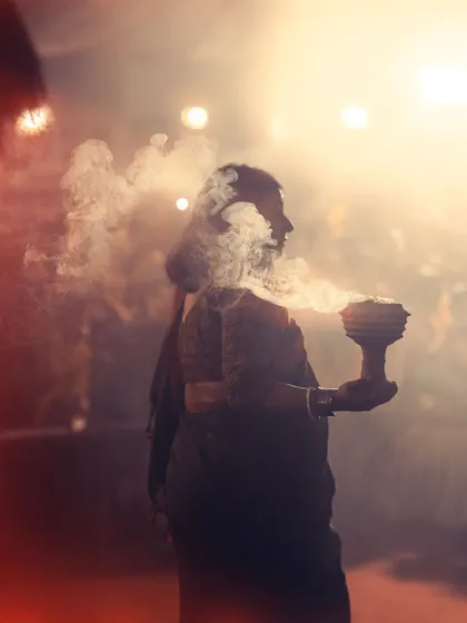 A silhouette of a woman holding a dhunuchi against the bright lights of the pandal. The smoke creates a beautiful, mystical effect around her.