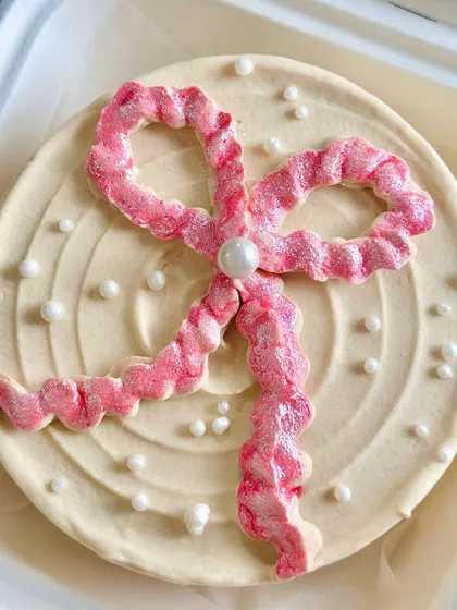 A close-up of a coquette-style bento cake, featuring a large, glittery pink bow made of buttercream and delicate pearl sprinkles.