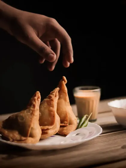 A food photography shot where a hand is reaching for a samosa, adding a human element and a sense of action to the scene.