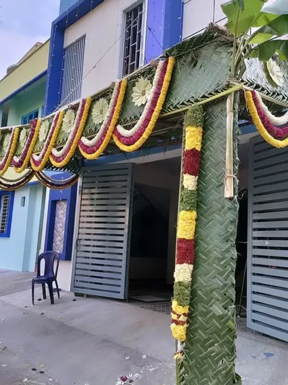 A wide chapra design covering the entrance gate of a home. The garlands of yellow, red, and white flowers add a vibrant touch to the green palm leaf structure.
