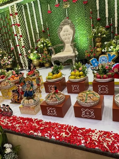 A traditional platter setup for a baby's naming ceremony. The display is arranged on decorative brown boxes and features dolls, fruits, and a sign announcing "It's a BOY".