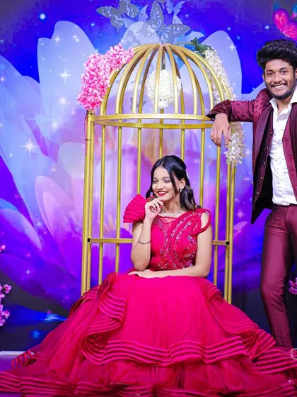 A whimsical studio shot of a couple with the woman in a tiered red gown seated in a golden birdcage prop, surrounded by a magical floral backdrop.