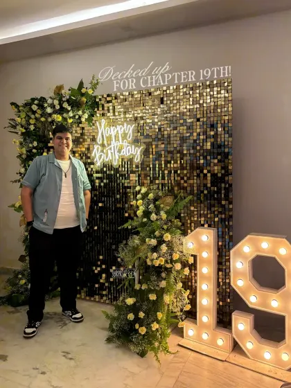 Decked out for chapter 19! The birthday boy, Pranav, posing in front of the elegant golden shimmer and floral backdrop we designed for his celebration.