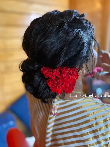 A soft, romantic low bun with wispy tendrils, decorated with a cluster of bright red carnations. This style is perfect for a rustic or outdoor wedding setting.
