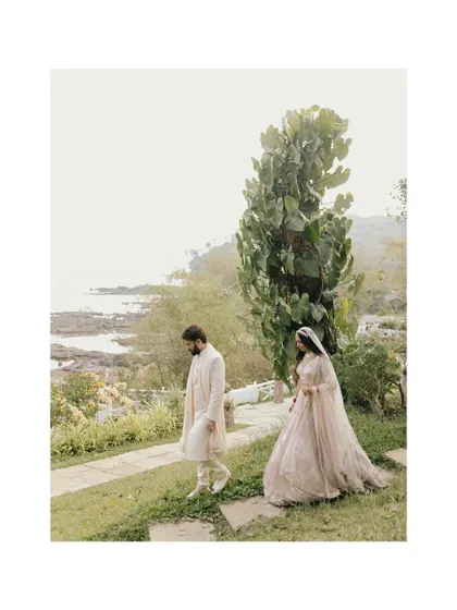 A serene, artistic shot of Amreet and Saksham walking in a coastal garden. The framing and soft focus create a dreamy, romantic feel.