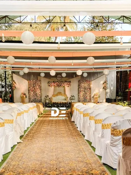 A daytime view of a semi-outdoor wedding ceremony setup. The aisle is lined with neatly covered chairs, leading to the stage, while paper lanterns hang from the ceiling for a touch of simple elegance.