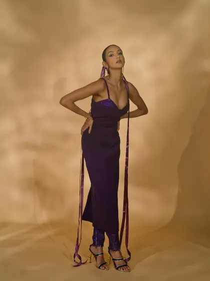 A full-length shot of a model in a purple gown with long ribbon details. The pose is dynamic, showcasing the unique design elements and modern feel of the outfit.