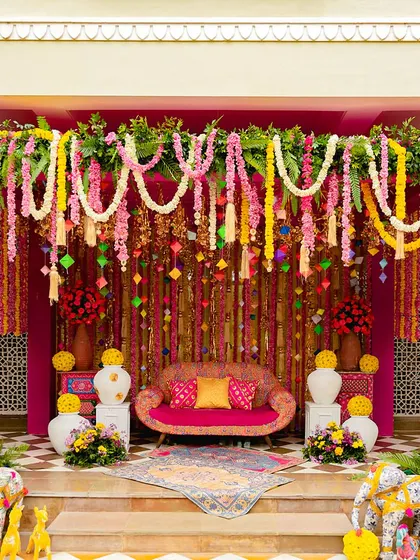 A vibrant and traditional bhaat ceremony. The seating area is a riot of pink and yellow, with floral garlands, colorful elephants, and a beautifully decorated sofa.