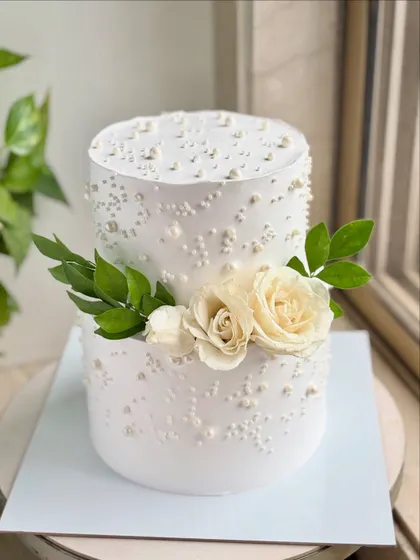 A classic two-tier wedding cake with a clean, modern aesthetic. The smooth buttercream is decorated with scattered pearls and a simple arrangement of fresh white roses and green leaves.